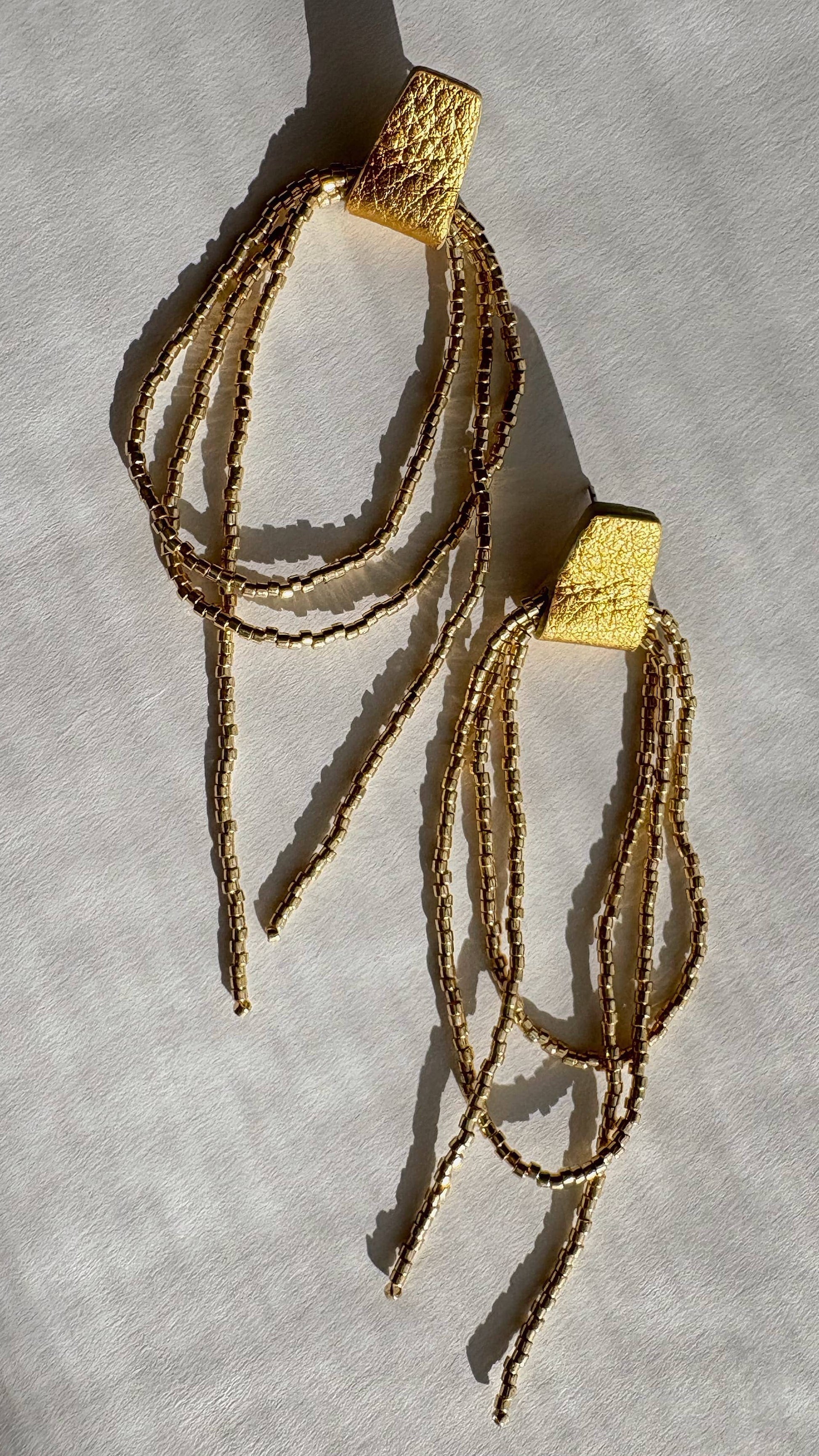 A pair of gold-toned statement earrings with textured square tops and multiple dangling beaded strands, displayed on a light fabric background with soft shadows.