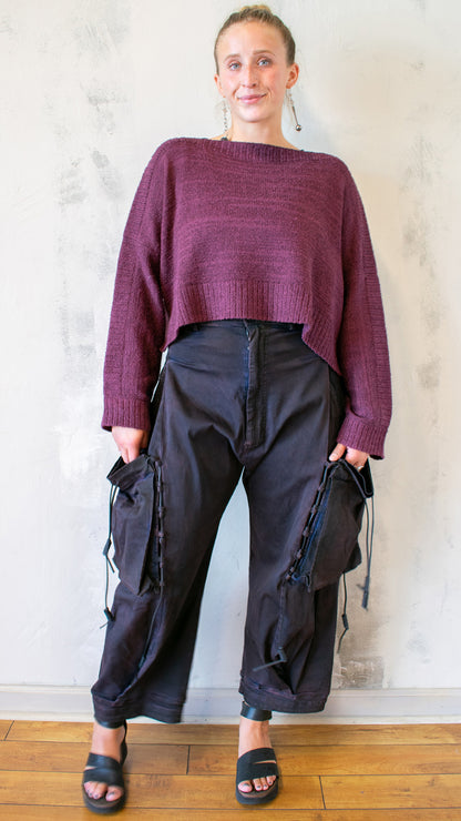 Person wearing a maroon sweater and dark pants standing against a light-colored wall.