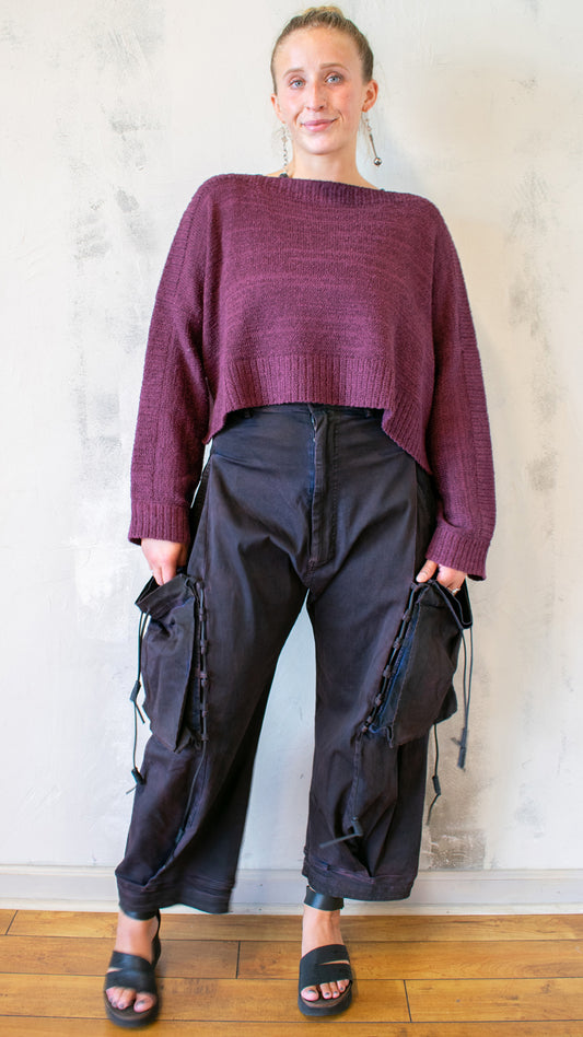 Person wearing a maroon sweater and dark pants standing against a light-colored wall.